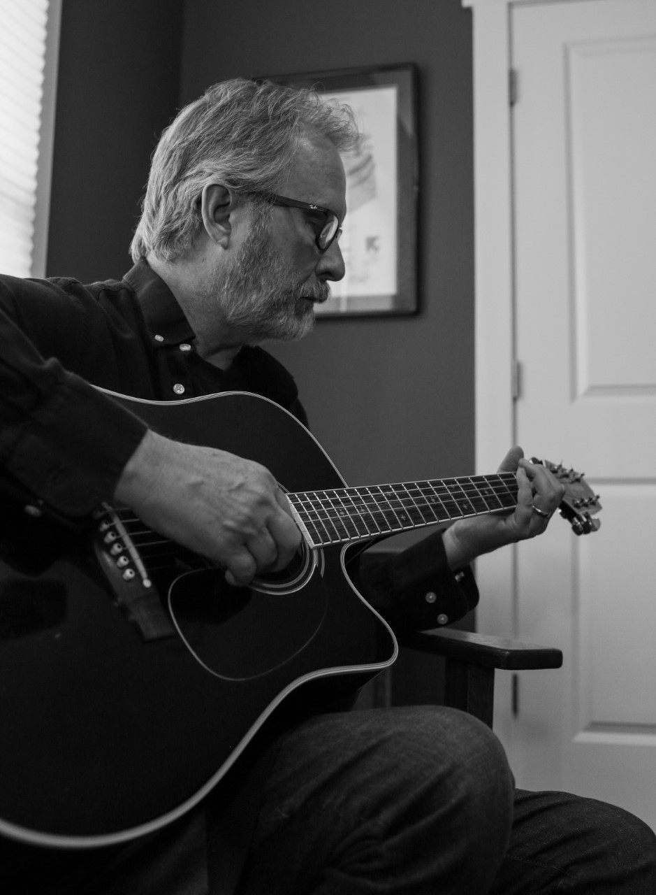 Black and white photo of Ron strumming a guitar while sitting. 