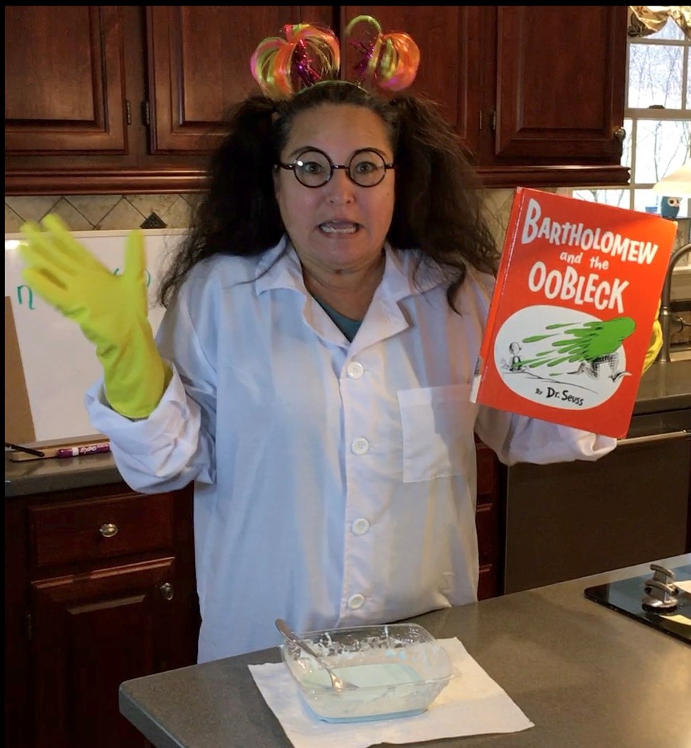 woman in labcoat yellow gloves holding Bartholemew and Oobleck book fluffy pigtails black round glasses orange and green headband sticking off