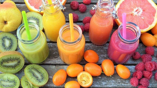 glass jars of gree, orange, and pink smoothies are surrounded by similar colored fruits and vegetables on a wood table