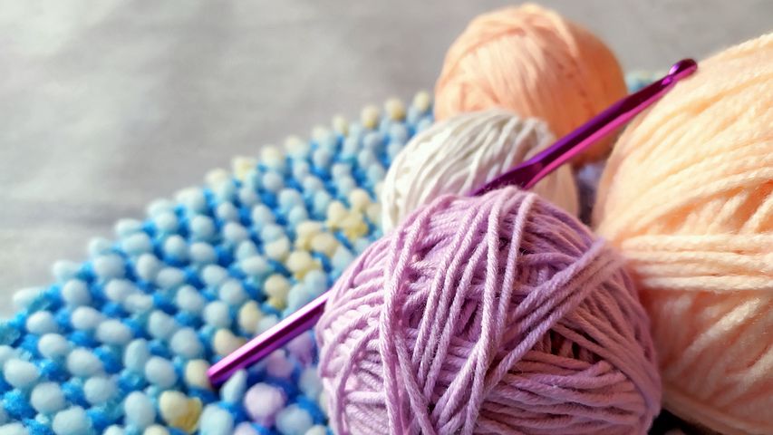 Pink, orange and white balls of yarn with a pink crochet hook sticking out - all laying on top of a a round edge of a blue crocheted item