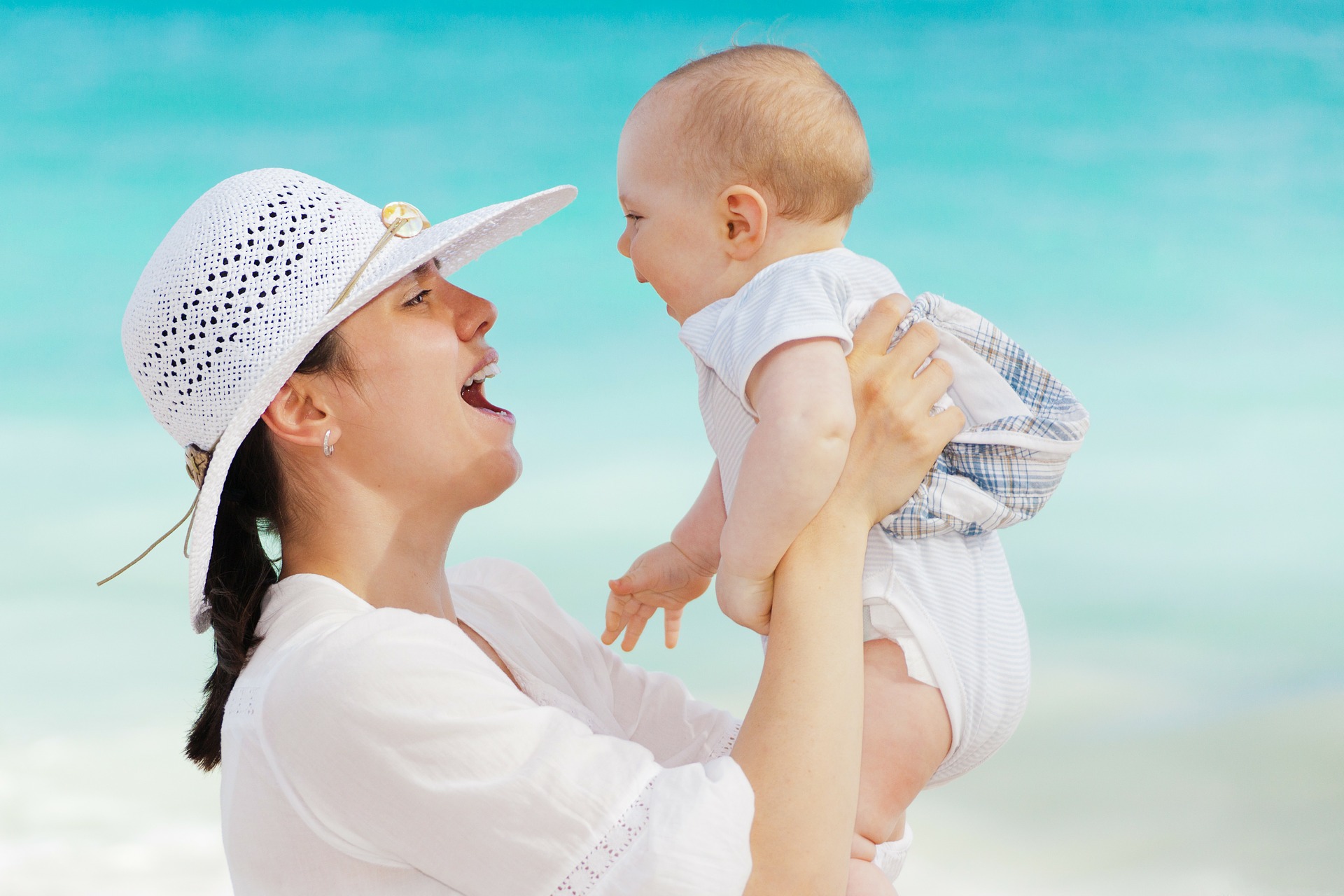 A Mother is holding baby up and smiling. They are at a beach.