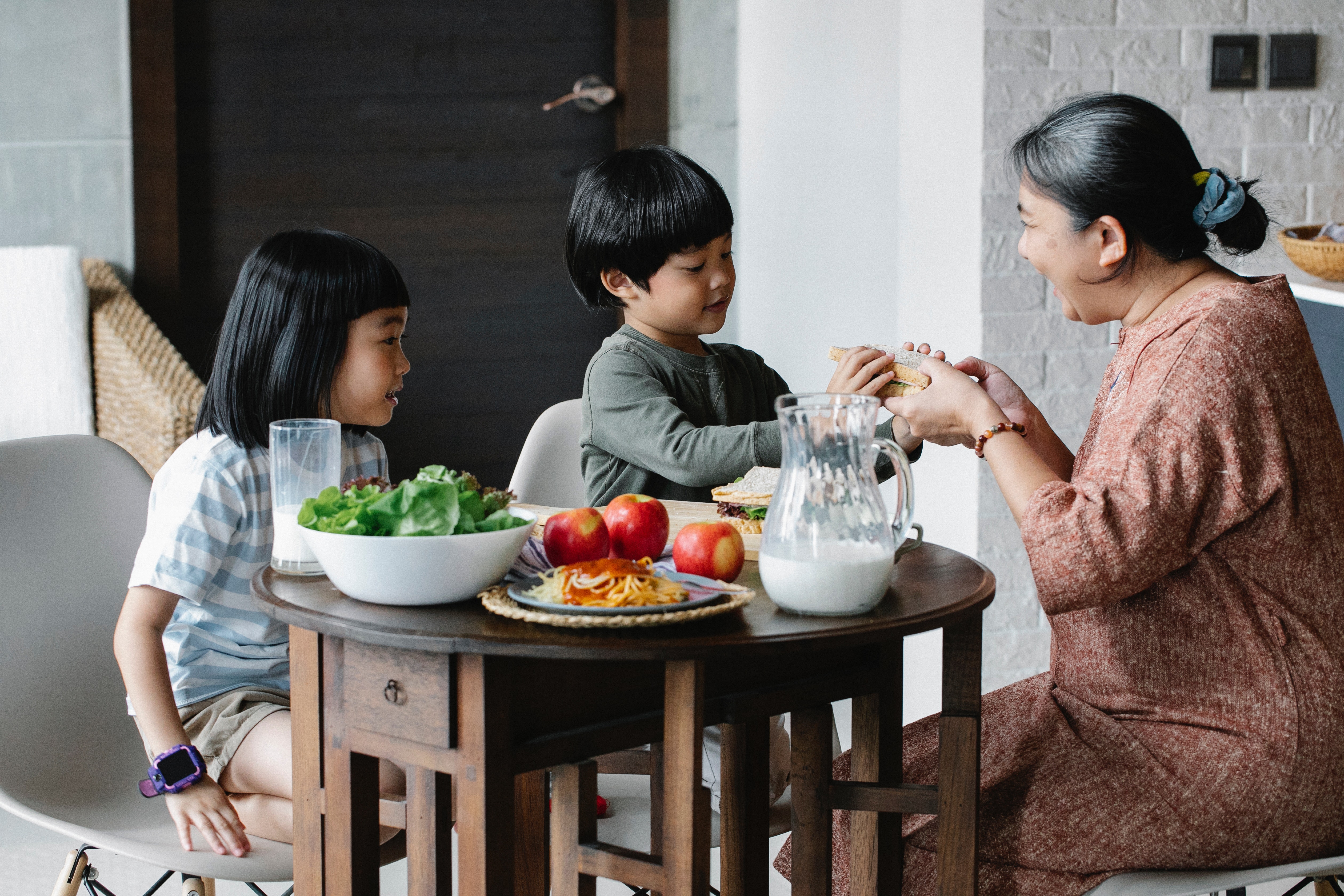 Photo by Alex Green from Pexels. A maternal figure sitting at a small table with two young children. On the table are a pitcher of milk, a bowl of lettuce and several apples. The adult is handing one of the children a sandwich.