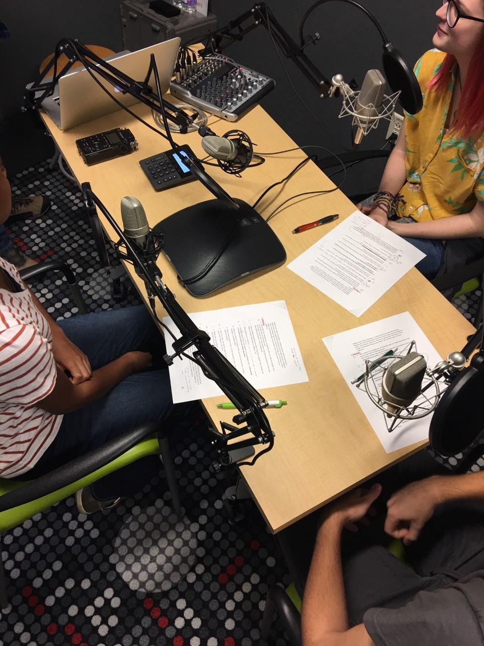 students sitting at a desk covered with papers and podcast recording equipment