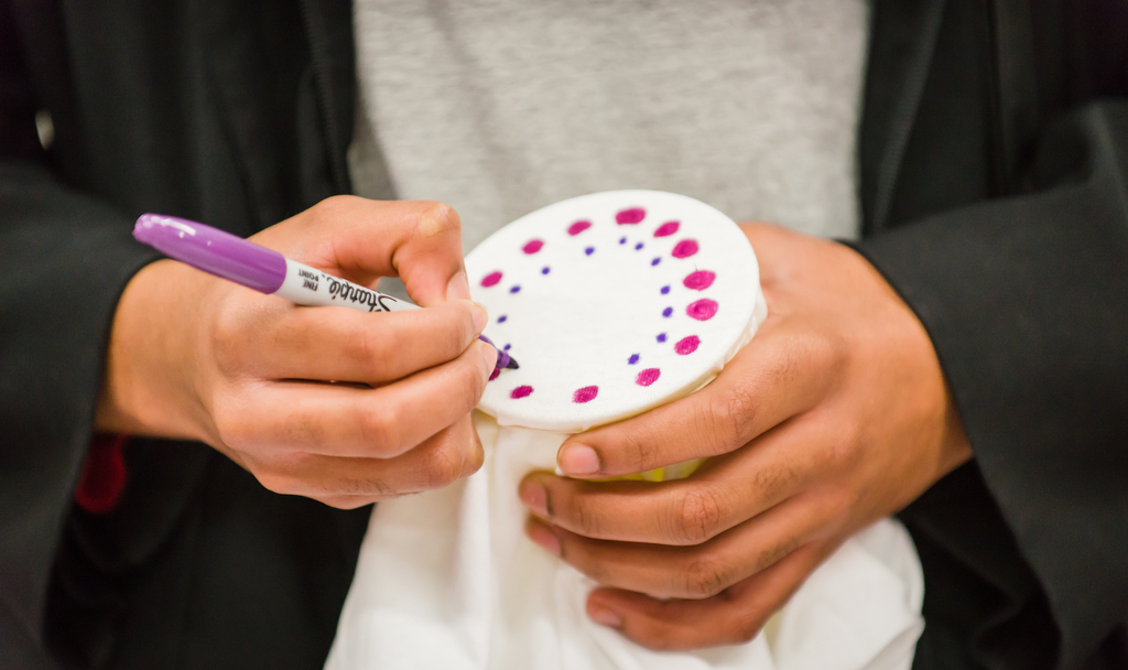 two hands holding a purple sharpie marker and drawing colored dots in a circular pattern on a white piece of fabric stretched over the top of a plastic cup