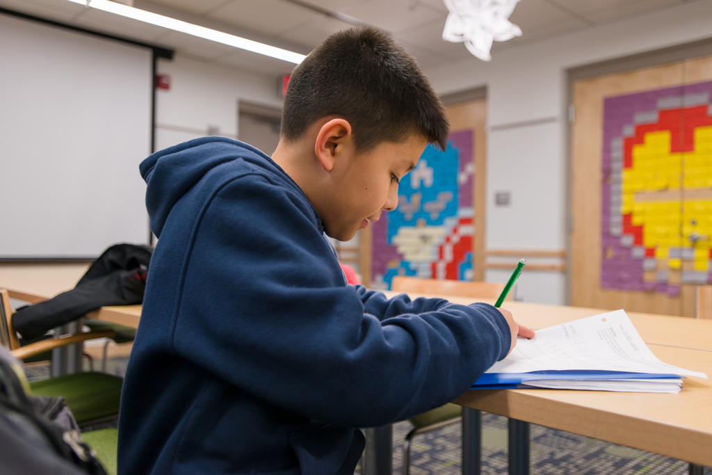 A child wearing a blue hoodie sits at a table writing on a paper