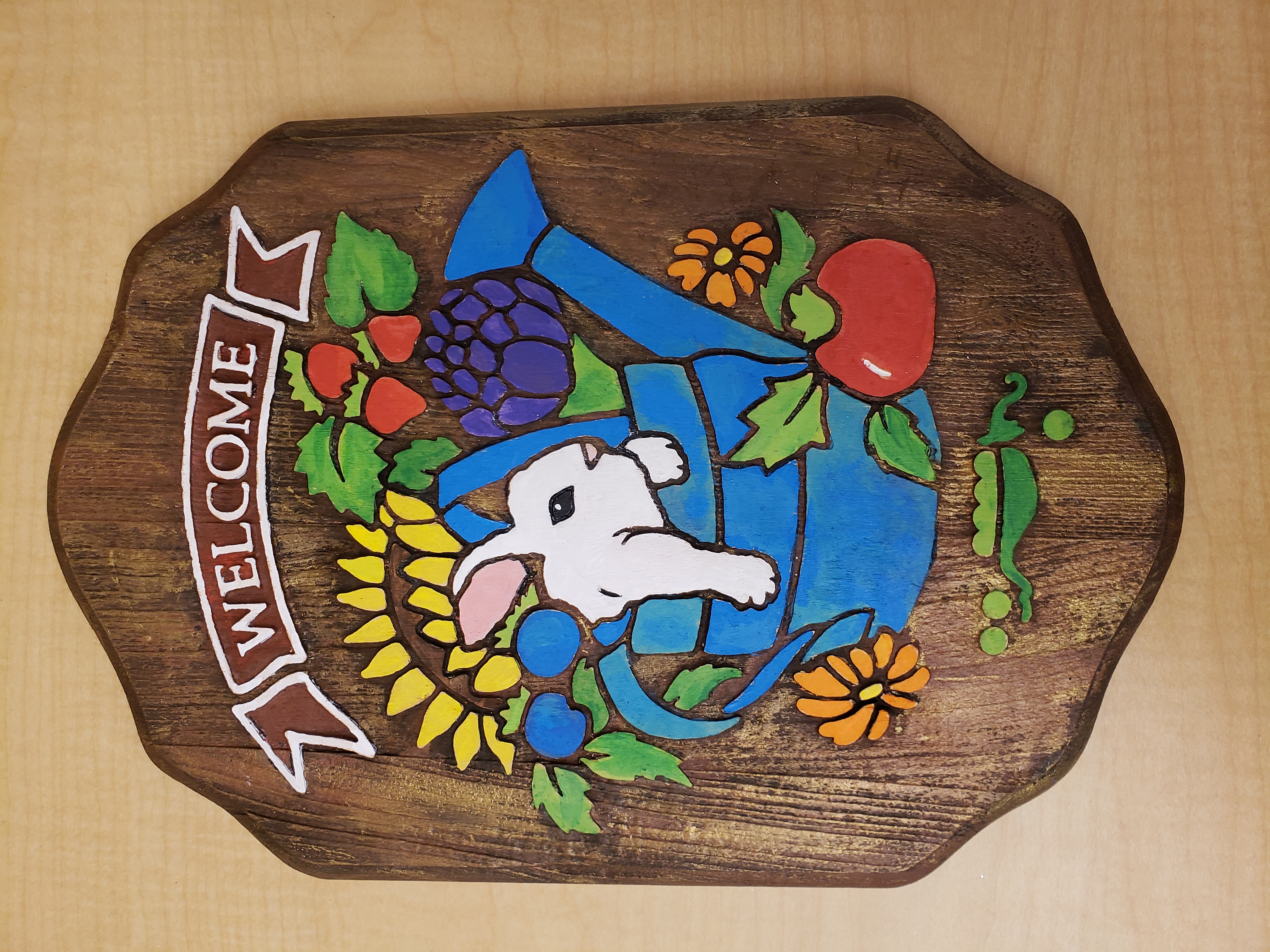 A painted wooden welcome sign featuring a rabbit in a watering can surrounded by plants and flowers