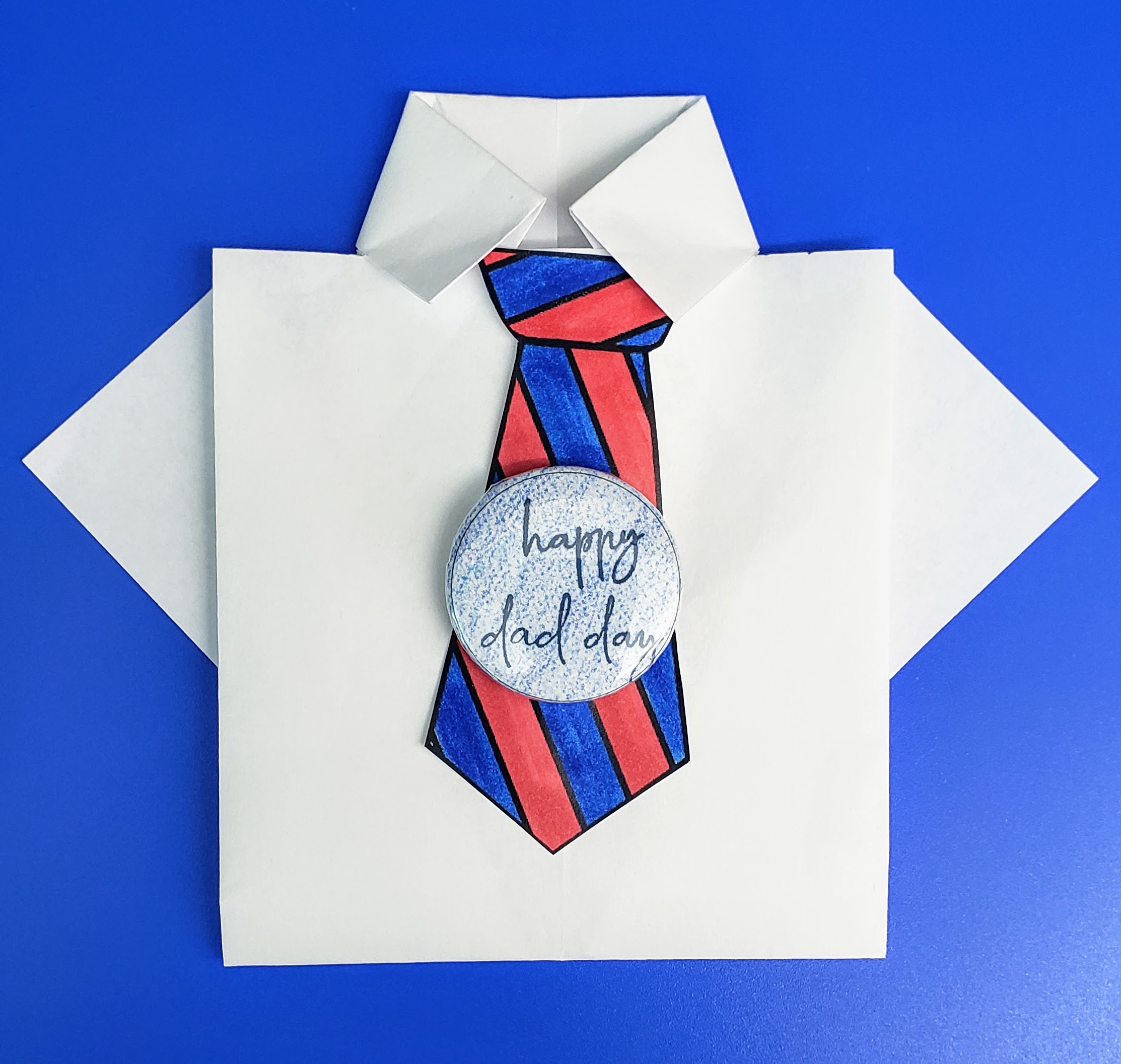 White origami paper shirt with blue and red striped paper tie. Button reading "happy dad day" on front of paper tie. 