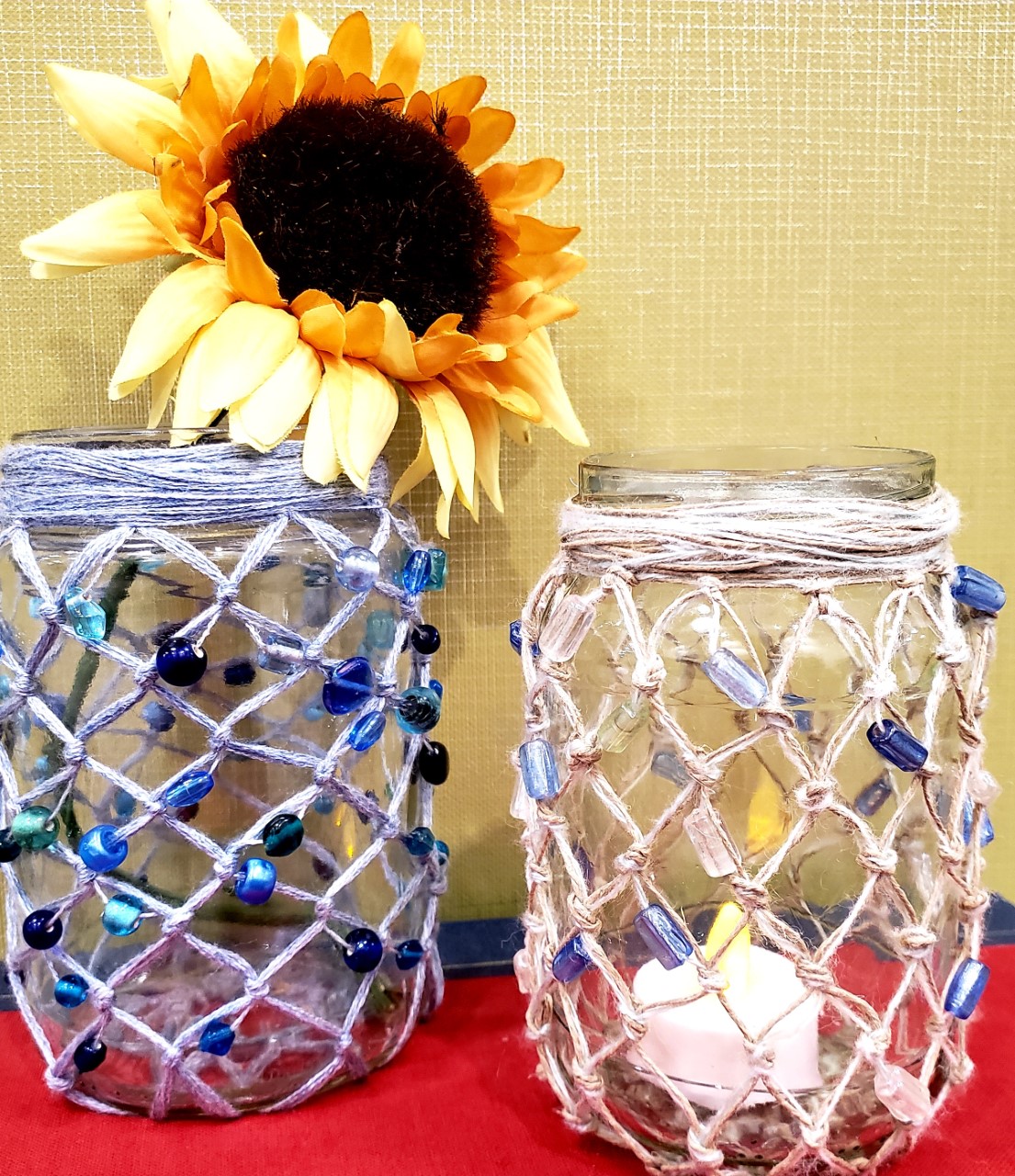 two jars covered in knotted and beaded string/yarn to create a net effect in one is a sunflower in the other is a small candle