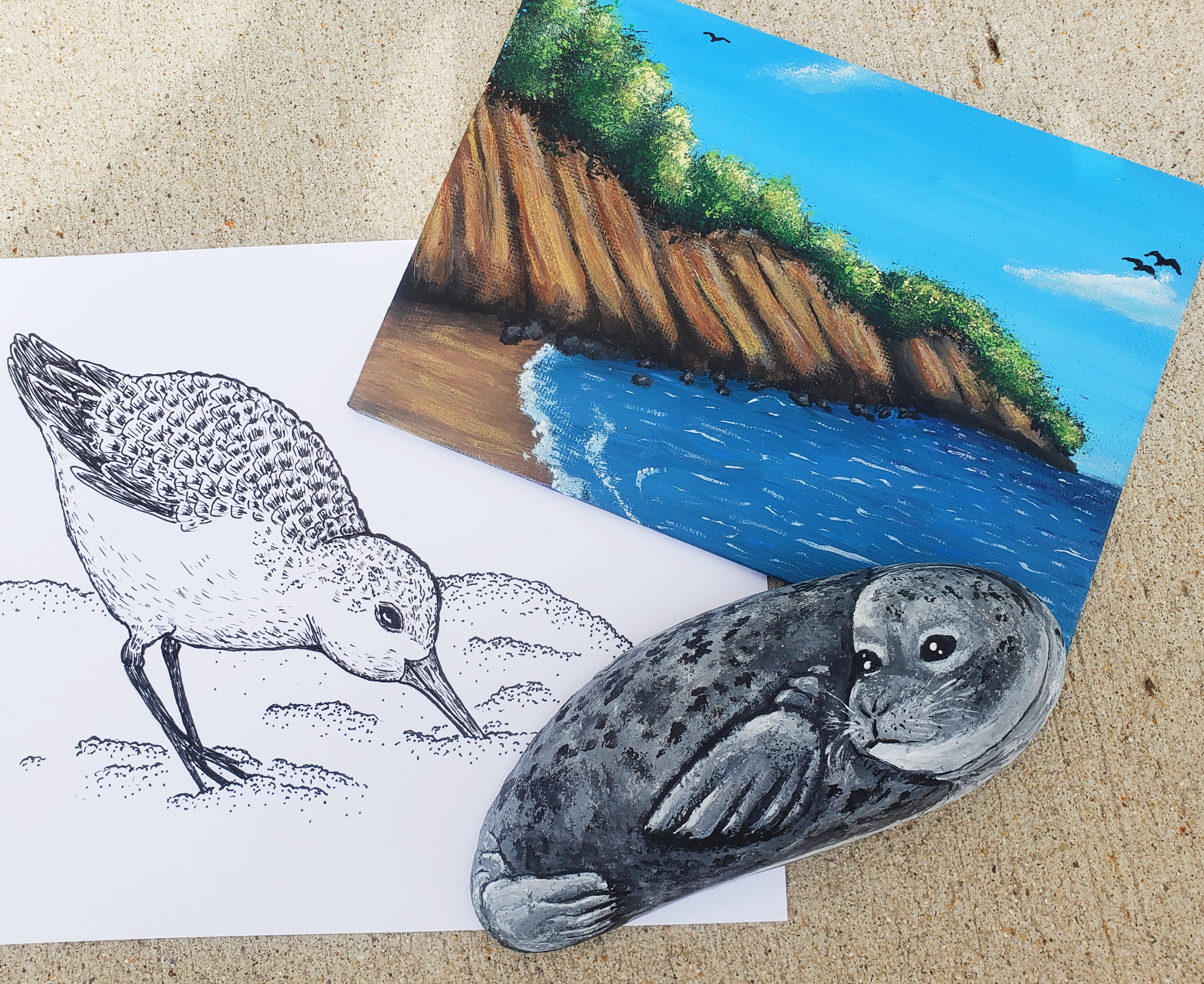 a pen sketch of  a sanderling (shore bird) a small painting of Calvert Cliffs beach and a rock painted to resemble a harbor seal