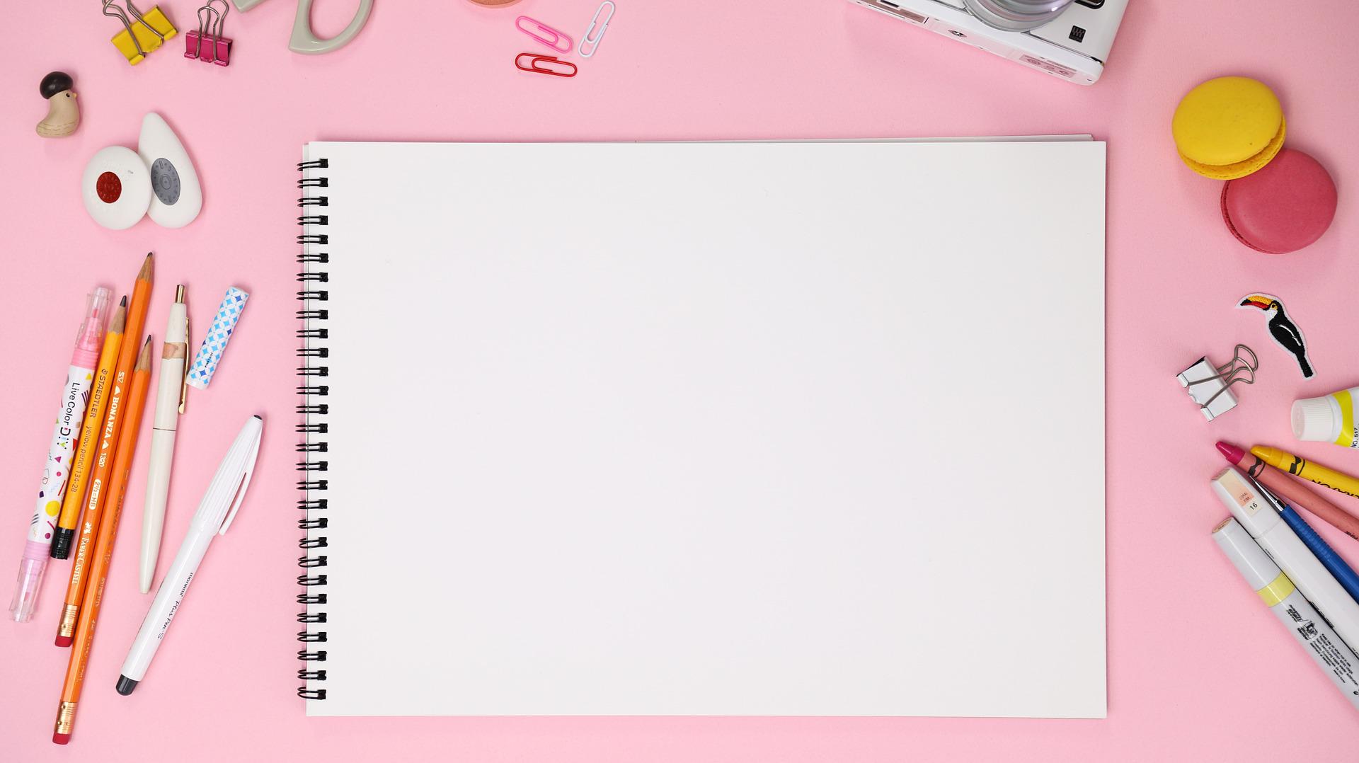 pink background with assorted pencils and drawing supplies with a drawing notebook opened to a blank page