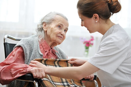 An older lady in a red shirt is in a chair and a nurse is covering her with a plaid blanket