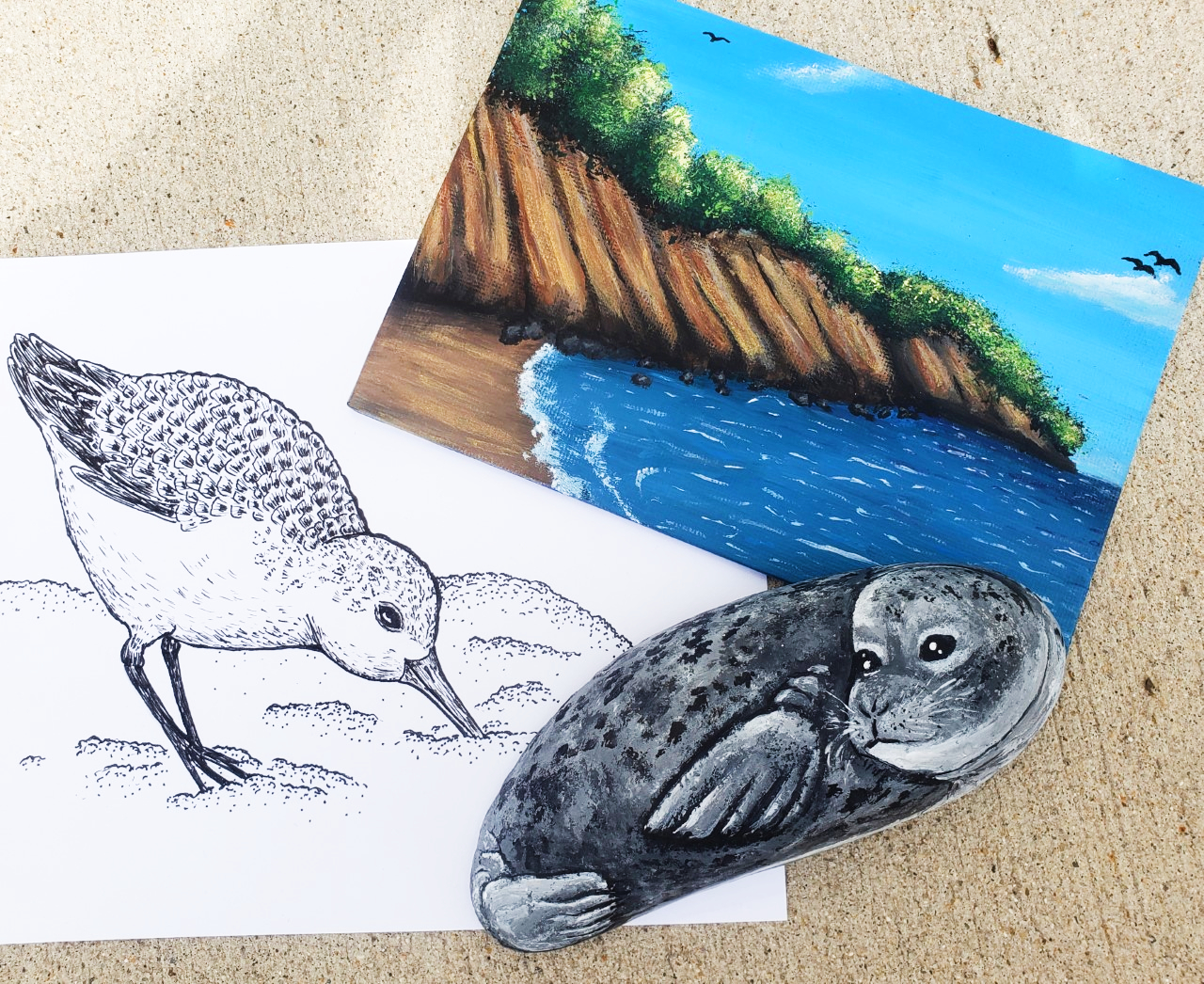 a painting of a beach, a pen drawing of a sanderling (bird) and a rock painted to look like a seal