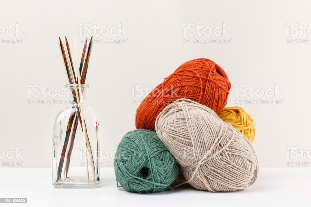 Multicolored bamboo wooden knitting needles in glass jar and red, green, yellow, and beige pastel colored yarn.