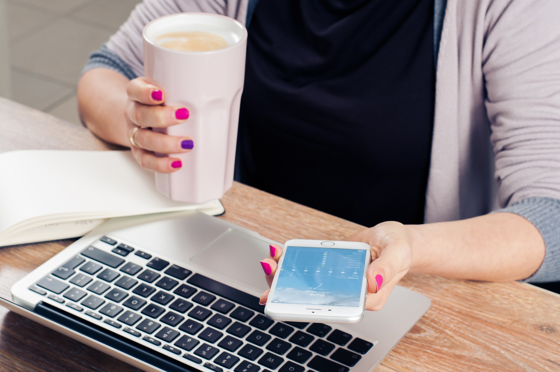 A person with red and purple painted nails sits at an open laptop while holding a cell phone and a cup of coffee
