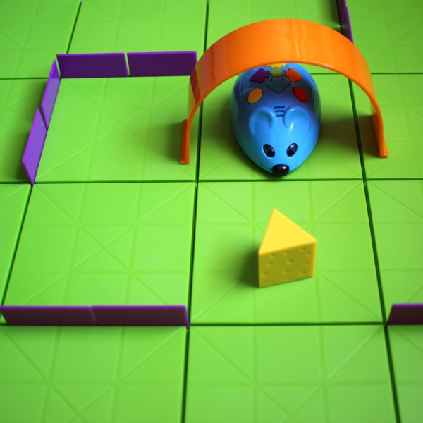 A blue robot mouse navigates maze of purple and orange pieces on a green board to reach a yellow wedge of cheese.