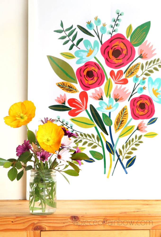 Paper flowers on a white background with a vase of different colored real flowers in a vase in front and to the left. 
