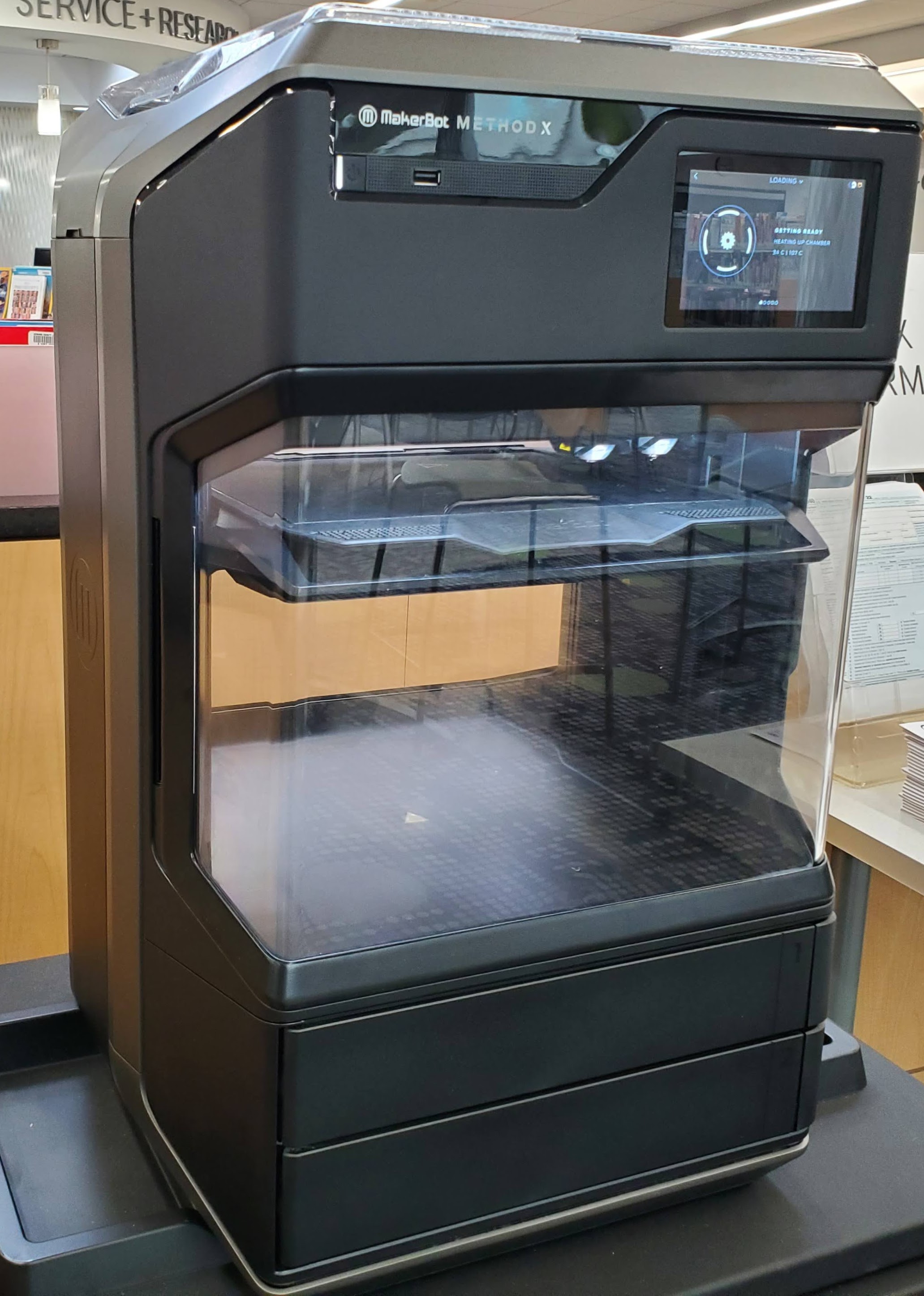 a 3D printer; a large dark grey plastic enclosure with a transparent door on the front, inside the print bed is raised and the extruders are visible near the top of the machine