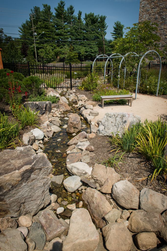a carefully landscaped garden: a rocky stream formed with stones is in the foreground, some mulch beds with plants surround it; moving away from the viewer is a bench, bushes, and a metal arbor. In the far back is a black metal fence.