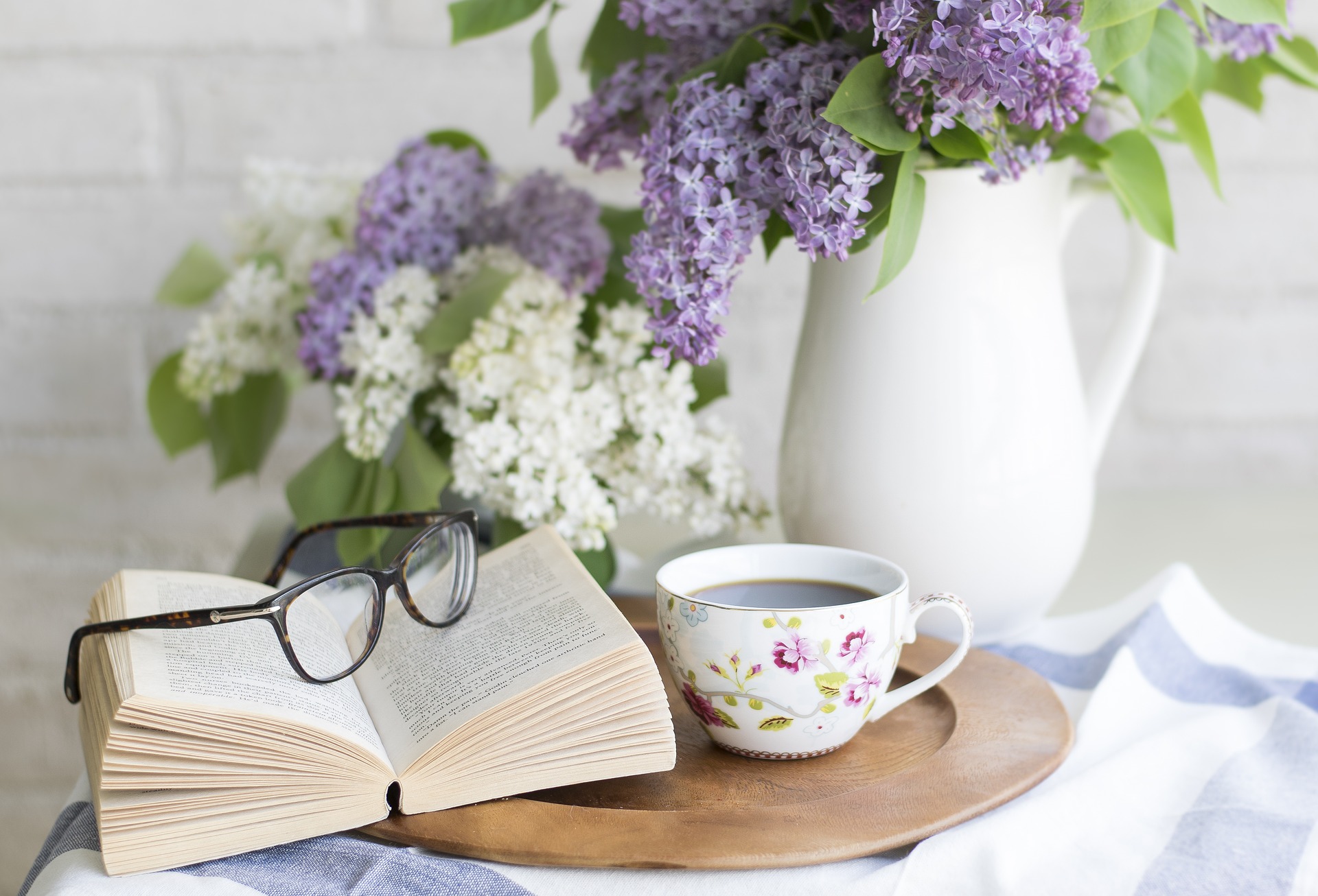 an open book with glasses on top and a white cup of coffee with a white vase with purple flowers