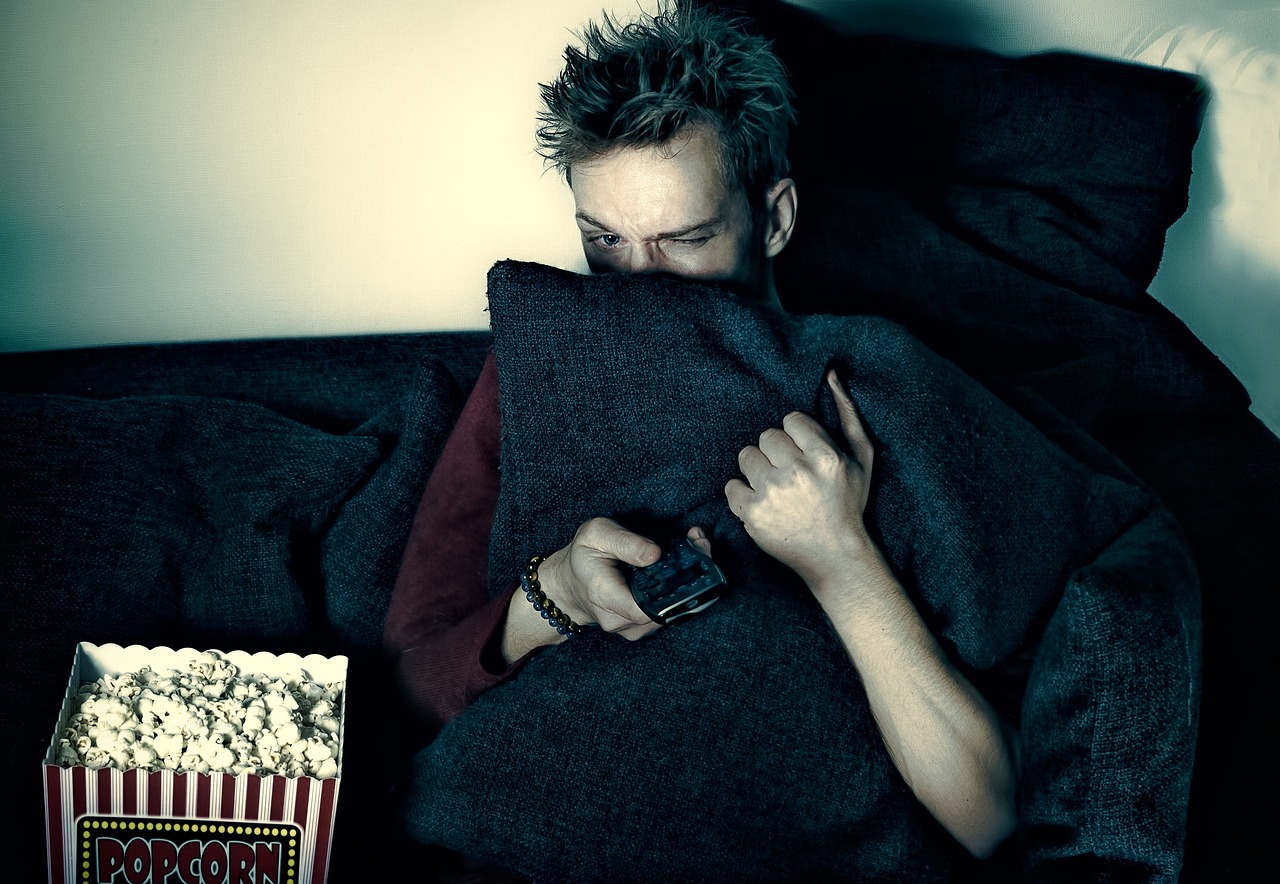 person watching tv with a bucket of popcorn, covering face partially with a pillow and holding the remote