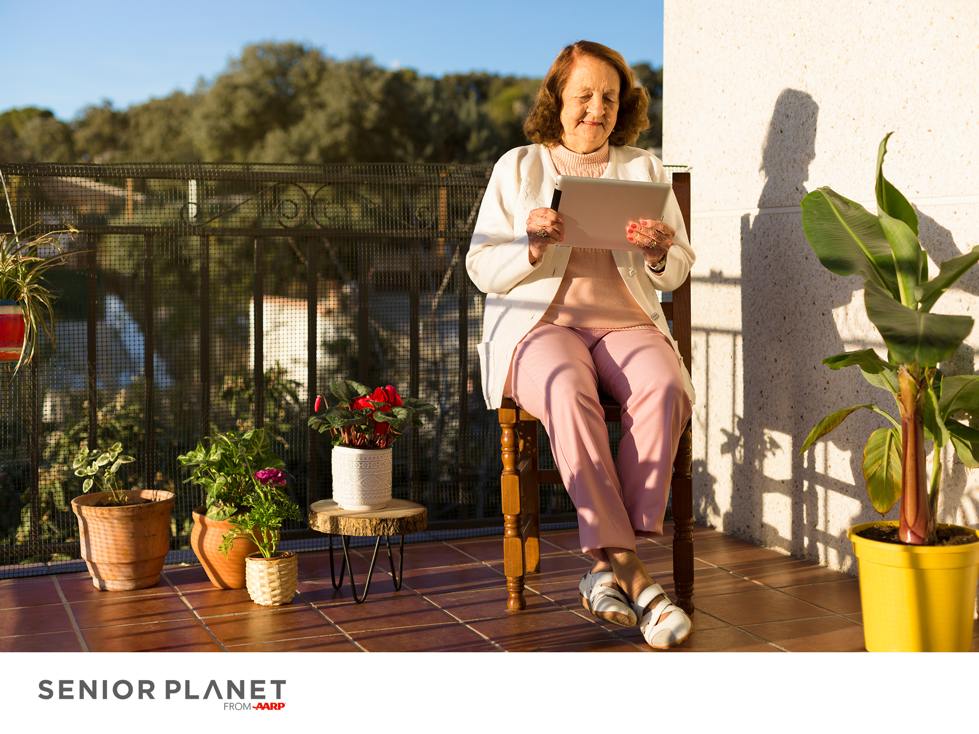 a smiling woman sitting in a chair on a balcony, holding a tablet computer and looking at it while smiling