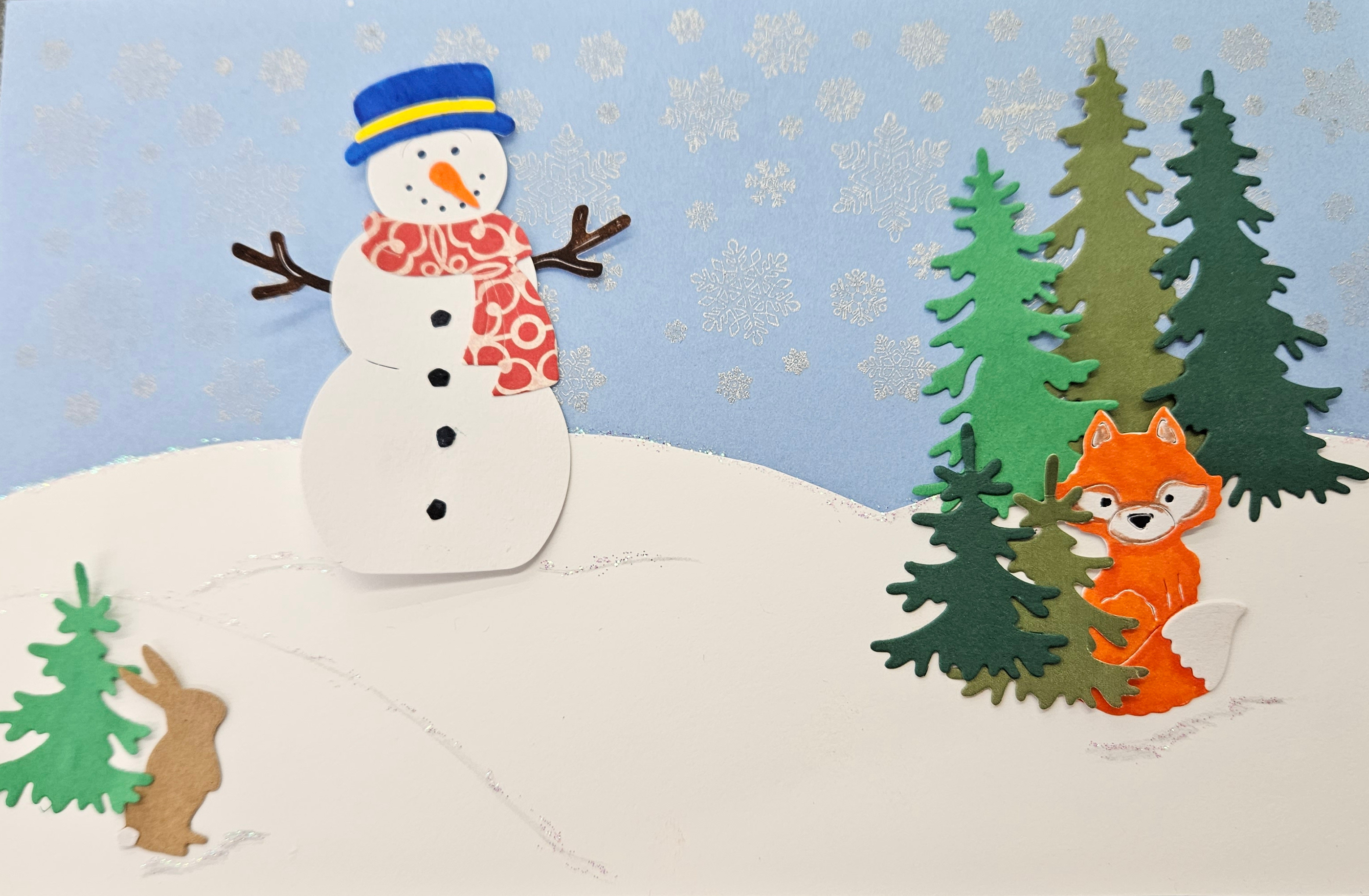 A winterscape scene made of colorful cardstock, featuring a snowman, green pine trees, a fox, and a rabbit. 
