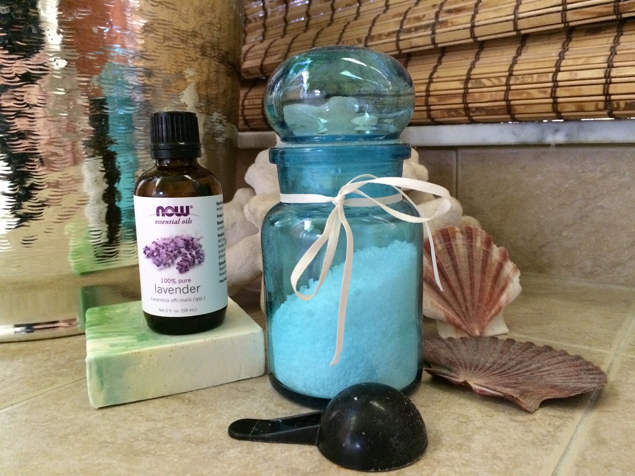 collection of beauty items on a tile counter, bottle of lavender essential oil on top of a block of soap, a jar of granular material, a tablespoon scoop, and some seashells