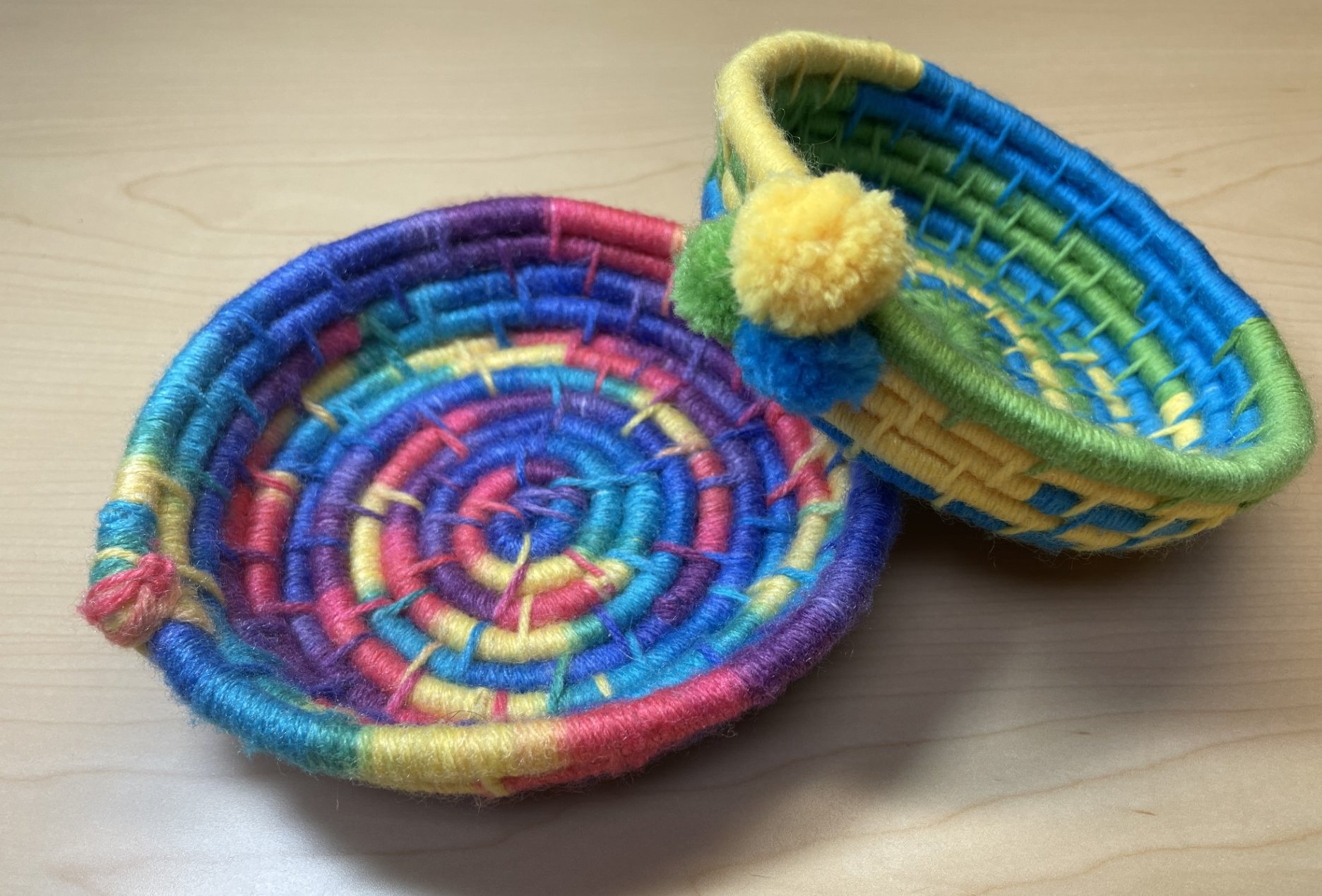 Examples of two woven yarn bowls. One rainbow colored, the other yellow, blue, and green with pom poms.