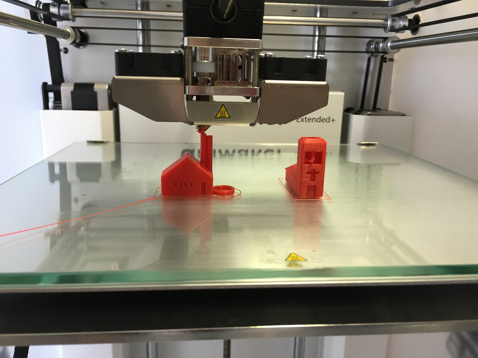 close up view of a plexiglass print bed, the metal gantry of a 3d printer can be seen surrounding, two orange house-shaped models are being printed, the extruder is visible completing printing of one of the houses