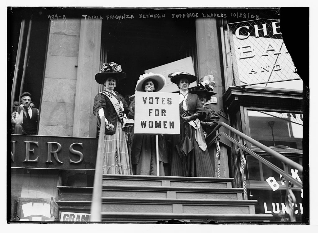 black and white photo, three suffragettes standing atop a staircase holding a sign reading Votes for Women