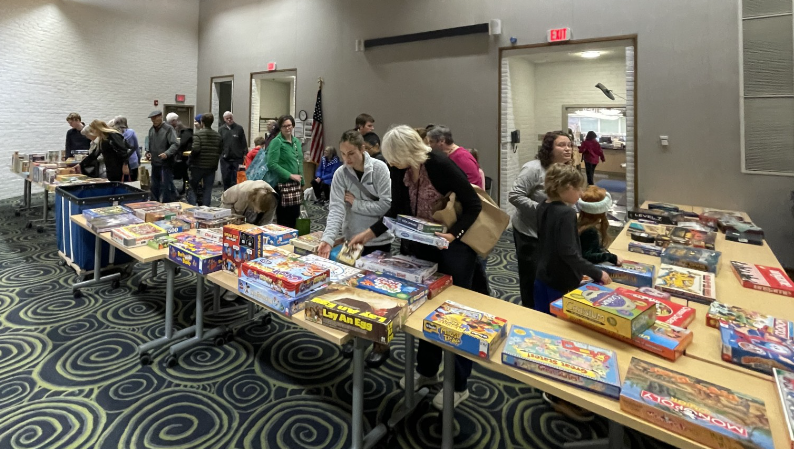 A group of people look at a variety of board games and puzzles laid out on tables. 