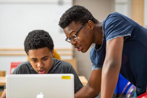 A person works at a laptop while another person leans over from the side to look at the same laptop.