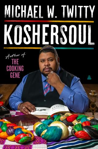 Michael Twitty is wearing a blue shirt and black vest, kippa, and sits on a table with dishes of food spread out 