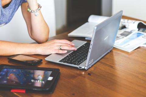 Cropped photo of a person using a laptop computer, a pile of papers sits to the left of the laptop and a tablet to the right