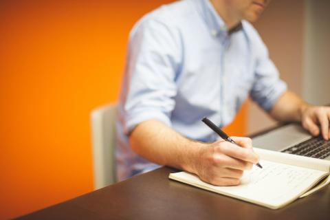 a man in a white shirt writing on a paper. Orange wall in the background