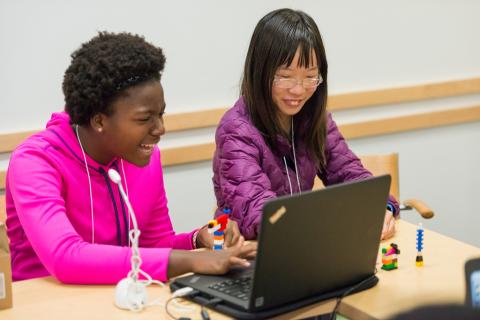 two teens laughing in front of a laptop computer screen with lego blocks on the table