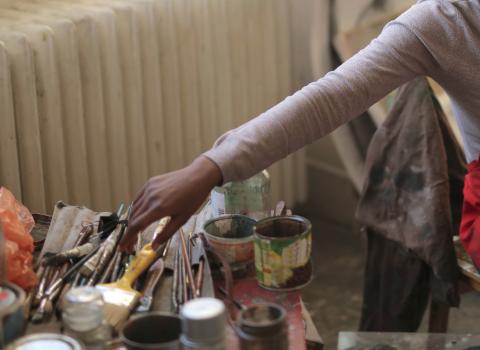 The arm of a Black painter stretches across the photo, reaching for a paintbrush. The painter has dark skin, and is wearing a light grey long-sleeve shirt and a red apron.