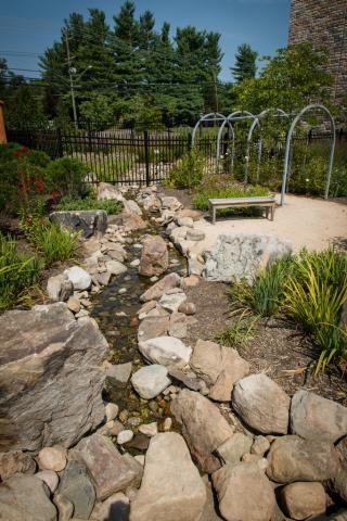 a carefully landscaped garden: a rocky stream formed with stones is in the foreground, some mulch beds with plants surround it; moving away from the viewer is a bench, bushes, and a metal arbor. In the far back is a black metal fence.