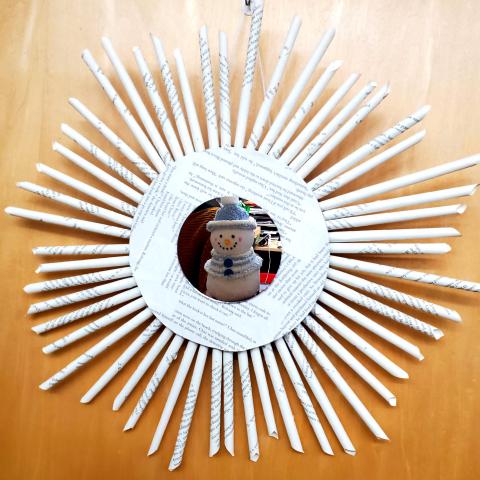 A cylindrical wreath made from rolled book pages, cardboard, and a circular mirror in the middle reflecting a snowman.