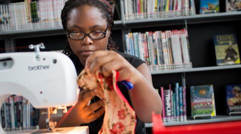 teenager holding fabric next to a sewing machine in front of a background of book shelves