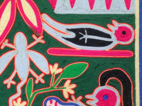 A close-up detail of a Huichol yarn painting. The image shows intricate patterns created with colorful yarn pressed into a surface. Vibrant colors including pink, multiple shades of green, white, blue, and red form geometric shapes and nature-inspired designs. Individual yarn strands are visible.