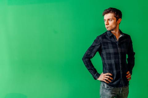 Man in a flannel shirt stands in front of a green backdrop with his hands on his hips