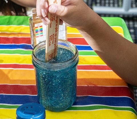 A person stirs blue glitter glue in a jar with a popsicle stick on a striped tablecloth.