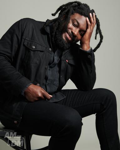 Photo of author Jason Reynolds sitting in a chair, smiling with his left hand to his face