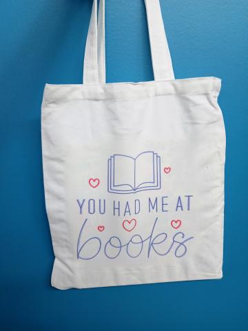 White tote back with red and purple writing. Shows a drawing of a book surrounded my hearts and reads, "You had me at books."