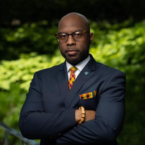 Headshot image of Marcus Nicks. Standing with arms folded. Wearing glasses, suit and tie. 