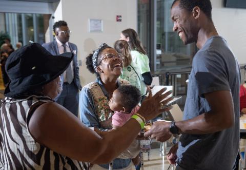 A Black woman holding a baby and laughing, people around her are smiling.