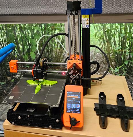 A Prusa Mini+ 3D printer using light green silk textured filament to 3D print an object.