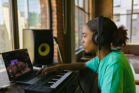 A woman wearing headphones sits in front of a laptop. Her computer has audio editing software open and a MIDI keyboard connected to it.