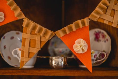 four pieces of pie made from felt strung together to create a garland. two slices of pie are pumpkin pie with a whipped cream topping, the other two are apple pie with criss-cross pie dough. 